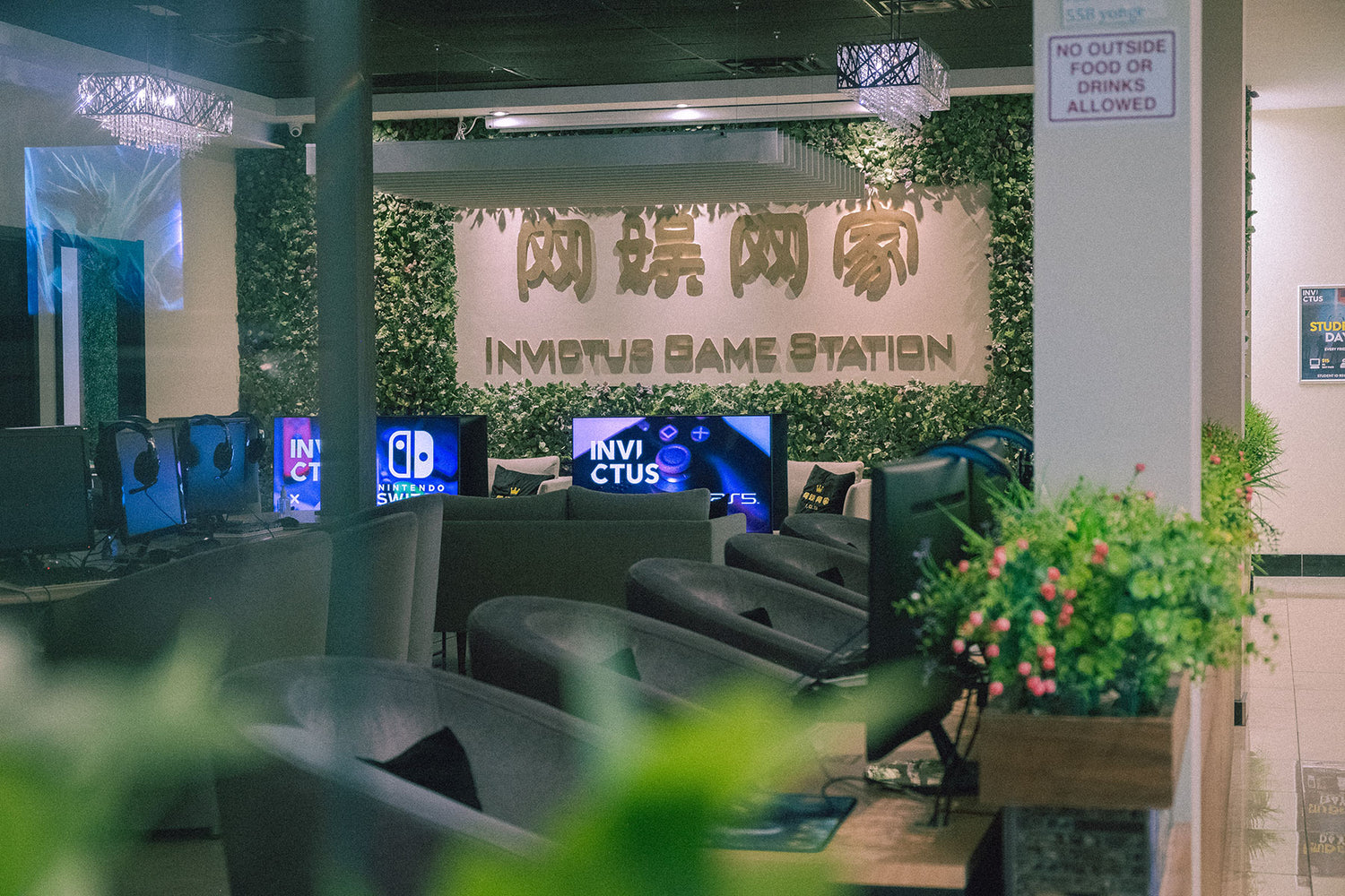 Inside a gaming cafe, several players are focused on monitors displaying a first-person shooter game. Foregrounded by out-of-focus green foliage, the screens show detailed in-game graphics, while in the background, a promotional gaming poster adds to the immersive environment.