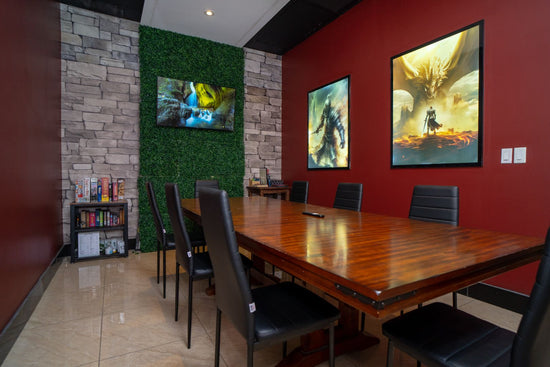A modern meeting room featuring a glossy wooden table surrounded by black chairs. One wall is accented with gray stone and adorned with a flat-screen TV displaying a nature scene, while the opposite wall is painted red and showcases fantasy-themed artwork. A bookshelf with board games and books adds a casual touch to the space.