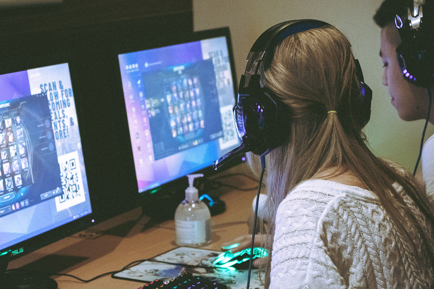 Side profile of a focused female gamer with long hair wearing a headset, sitting in front of a computer screen displaying a character selection menu for a video game. A QR code for gaming deals and a hand sanitizer bottle are also visible on the desk, indicating health safety measures and interactive promotions in the gaming space.