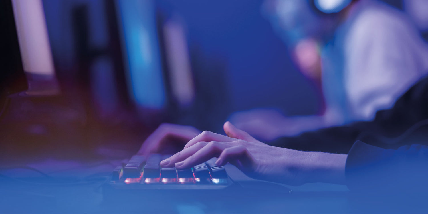 Close-up of a gamer's hands expertly navigating a backlit mechanical keyboard, with a blurred background showing another player wearing headphones, immersed in gameplay. The blue lighting casts a cool glow, emphasizing the intense focus and high-tech environment of competitive gaming.